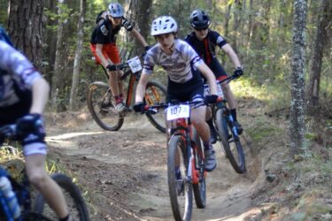 A group of young mountain bikers navigate a dirt trail through a wooded area. The trail features twists and turns, with trees visible in the background. One rider, wearing a white helmet and jersey, is in the foreground, while two other cyclists follow closely behind on the path. Mt. Zion Bike Trails mountain bike trail.