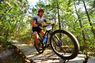 A mountain biker navigating a wooden bridge in a forested area, with tall trees and greenery surrounding the trail. The cyclist is wearing a red helmet and cycling gear, focused on maintaining speed and balance. Mt. Zion Bike Trails mountain bike trail.