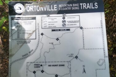 Map sign for Ortonville Mountain Bike Trails and Cross-Country Skiing, featuring trail markers, key for navigation, and notes regarding park rules and zones, surrounded by trees and foliage. Ortonville trail mountain bike trail.