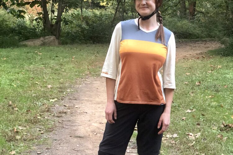 A person standing on a dirt path in a wooded area, wearing a black bicycle helmet and outdoor athletic attire, including a multi-colored shirt and shorts. They have a friendly expression and are posed with one hand on their hip. The surrounding environment includes trees and grass, with fallen leaves on the ground.