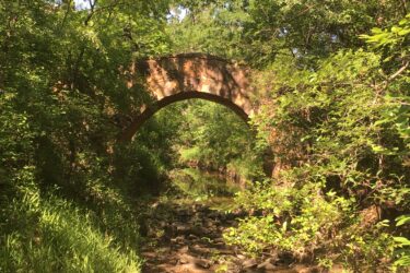 A stone arch bridge partially obscured by lush green foliage, spanning a shallow stream with pebbly banks. Sunlight filters through the trees, casting dappled light on the scene. Western Heritage Park mountain bike trail.