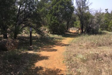 A dirt trail winding through a lush, green landscape with dense trees on either side. A wooden signpost indicating trail directions stands on the left, while a red bicycle rests against a tree in the distance. The ground is visible with patches of grass and dirt under bright, sunny skies. Western Heritage Park mountain bike trail.
