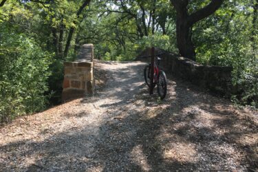 A gravel path leading over a small stone bridge, surrounded by lush greenery and trees. A mountain bike is parked on the side of the path, indicating a peaceful outdoor setting ideal for biking or exploring nature. Western Heritage Park mountain bike trail.