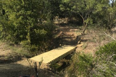 A wooden bridge crossing a small ravine in a natural landscape, surrounded by greenery and trees, with a mountain bike resting on the ground nearby. Western Heritage Park mountain bike trail.