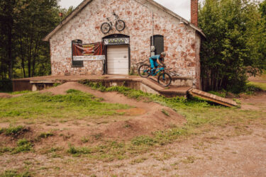 A mountain biker wearing a blue outfit performs a jump on a ramp in front of a rustic building with stone walls, which has bicycles displayed on it. Surrounding the area are patches of green grass and dirt jumps, indicating a biking park or trail. Banners are visible on the building, featuring the names of biking organizations. RAMBA Trails mountain bike trail.