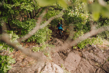 A mountain biker navigates a rocky trail through dense greenery in a forested area. The scene captures the cyclist from an elevated perspective, showcasing the challenging terrain surrounded by lush leaves and trees. RAMBA Trails mountain bike trail.
