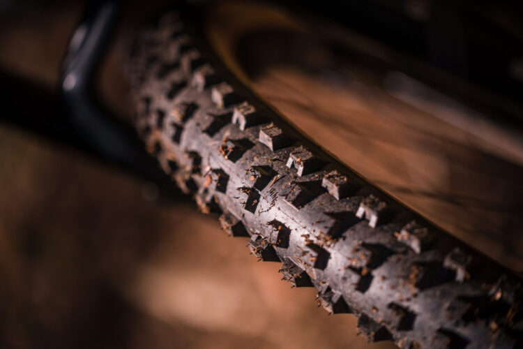 Close-up image of a mountain bike tire featuring pronounced, rugged treads with some dirt and debris, against a blurred background.