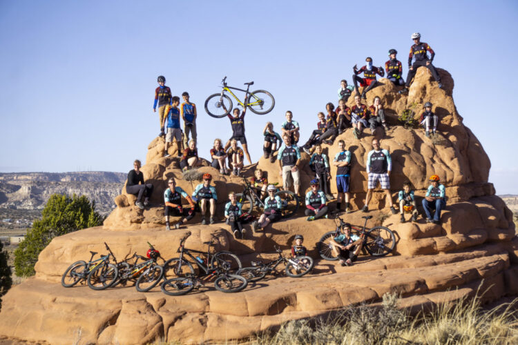 A large group of people posing on a rocky outcrop, celebrating a mountain biking event. Some individuals are holding bicycles, while others are seated or standing on the rocks. The scene is set against a clear blue sky and a picturesque landscape in the background, showcasing the excitement and camaraderie of the biking community.