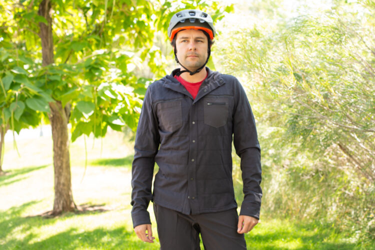 A man stands outdoors wearing a black long-sleeve shirt and a red t-shirt underneath, paired with a gray helmet. He is surrounded by lush greenery in a park setting, with trees and grass in the background. He appears to be prepared for an outdoor biking or recreational activity.