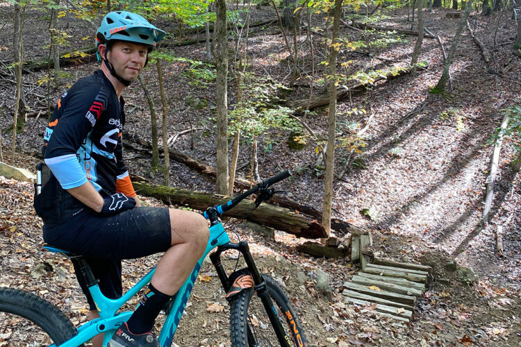 A mountain biker in a helmet sits on a blue bike, positioned beside a trail in a wooded area. The background features trees with autumn-colored leaves and a trail marked by wooden structures. The ground is covered in fallen leaves.