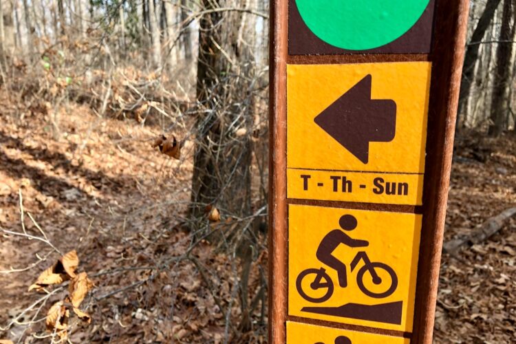 Trail sign indicating a bike trail. It features symbols for biking and directional arrows, with text indicating trail availability (Tuesday, Thursday, and Sunday). The background shows a wooded area with fallen leaves.