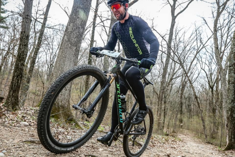 A mountain biker riding a black bicycle on a rocky trail in a forested area, wearing a black long-sleeve jersey with green accents and a helmet with tinted glasses. Trees without leaves fill the background, suggesting early spring or late autumn.