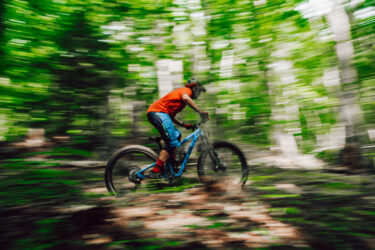 A young cyclist in an orange shirt rides a blue mountain bike through a green forest, captured in motion with a blurred background, conveying a sense of speed and adventure. Off Grade mountain bike trail.