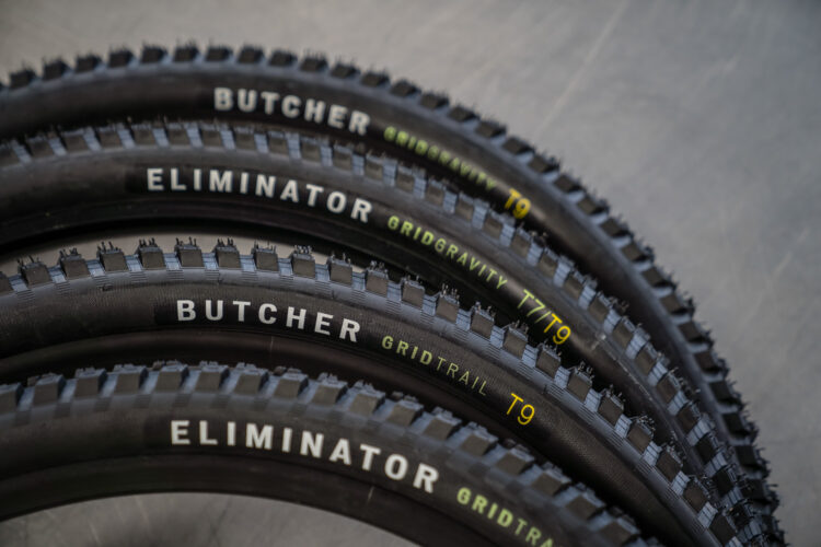 Four mountain bike tires arranged in two stacks, showcasing the tread pattern. The tires are labeled "Butcher" and "Eliminator," with markings indicating "Gridgravity," "Gridtrail," and various performance ratings (T7, T9). The background is a neutral gray surface.