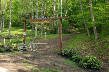 Sign marking the entrance to Eleanor Park Trails, with a mountain motif at the top. A mountain bike leans against the sign, and colorful flowers and lush greenery surround the entrance. A dirt path leads into a wooded area in the background. Eleanor Park mountain bike trail.
