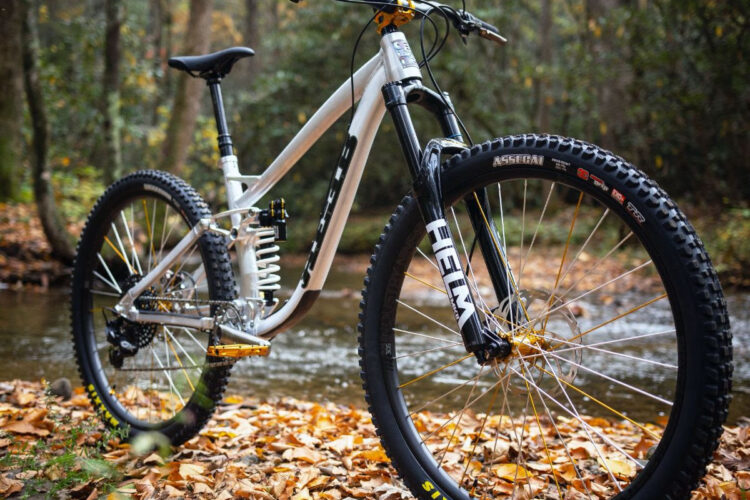 Mountain bike parked on a bed of autumn leaves near a creek, with trees in the background. The bike features a sleek white frame, black front fork, and wide tires designed for rugged terrain.
