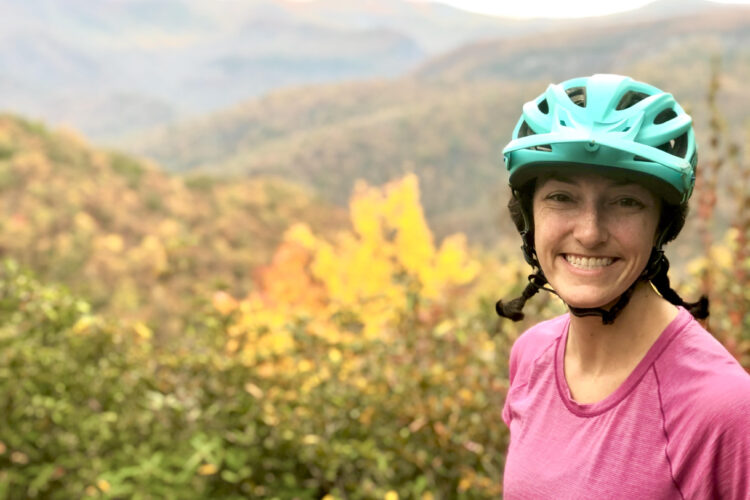 A woman wearing a turquoise helmet and a pink shirt smiles while standing in a scenic outdoor setting with colorful autumn foliage and mountains in the background.