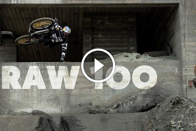 A BMX rider performing a trick off a ledge in an industrial setting, with "RAW 100" prominently displayed on the wall below. The scene captures the dynamic energy of extreme sports, showcasing the rider mid-air against a concrete backdrop.