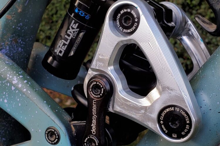 Image of a mountain bike suspension system, featuring a black shock absorber labeled "Super Deluxe" and a silver linkage component. The background is a blurred green landscape, highlighting the bike's intricate design and mechanics.