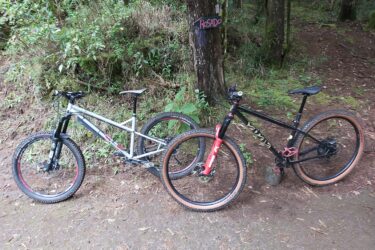 Two mountain bikes are parked on a dirt path surrounded by lush green foliage. The bike on the left is silver with a black front fork, while the bike on the right is black with red accents and a unique wider-tread tire. A wooden sign can be seen in the background, indicating a trail named "Rosado." Adventure Park mountain bike trail.