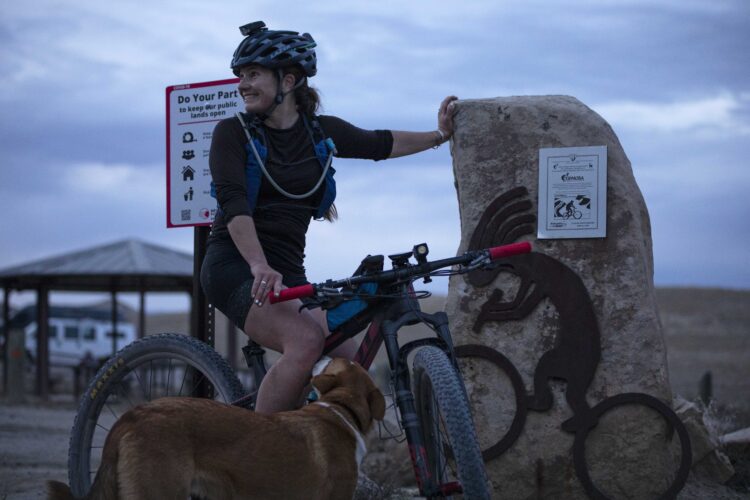 A woman wearing a helmet and cycling gear is sitting on her mountain bike next to a large rock with a bicycle-themed sculpture. She is smiling and appears to be interacting with her dog, which is nearby. A sign in the background encourages responsible use of public lands. The setting is outdoors, possibly during twilight, with a cloudy sky.