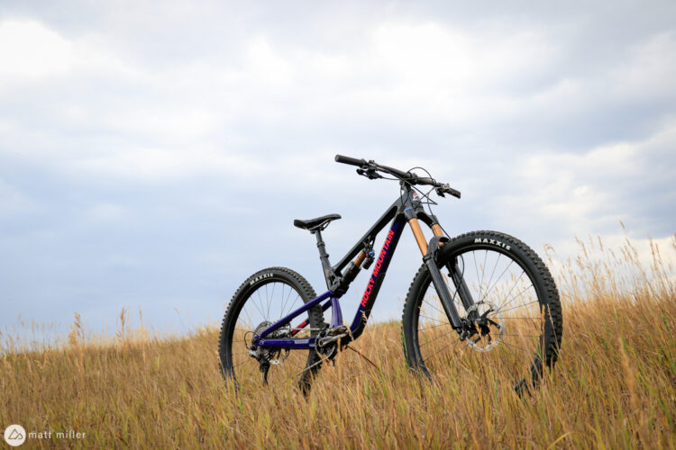 A mountain bike stands upright in a field of tall grass under a cloudy sky. The bike features a sleek black and purple design, with prominent branding visible on the frame.