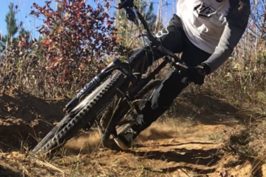 A person in a black helmet and protective gear rides a mountain bike on a dirt trail, leaning into a turn. Surrounding vegetation includes tall grass and bushes, with a clear blue sky above. Dust is kicked up from the bike tires, highlighting the action of the ride. Burning Love MTB Flow Trail mountain bike trail.