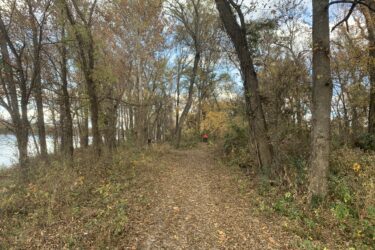 A tranquil forest path lined with trees displaying autumn foliage, leading towards a body of water. Leaves cover the ground, and a person in the distance walks along the trail. The sky is partly cloudy, creating a peaceful atmosphere. Three Forks Trail mountain bike trail.