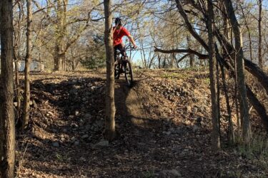 A person riding a mountain bike down a small dirt ramp surrounded by trees and rocks, with dry leaves on the ground in a natural setting. The rider is wearing a red shirt and a helmet, poised in mid-action as they navigate the trail. Three Forks Trail mountain bike trail.