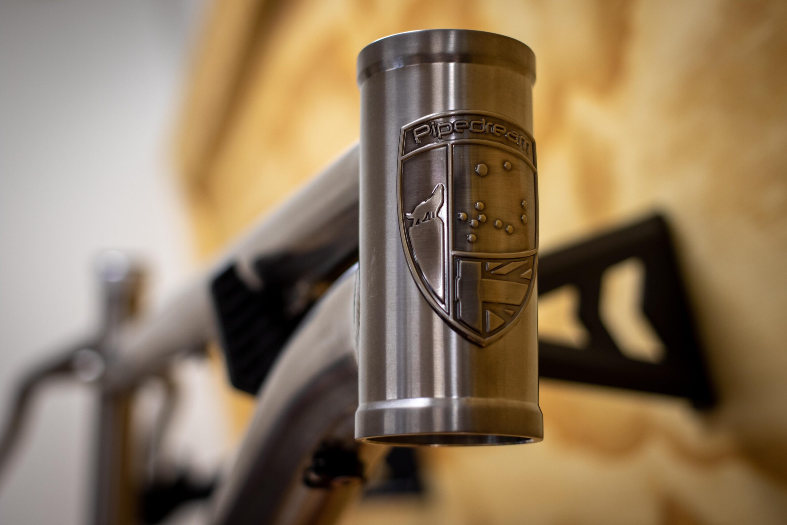 Close-up of a bicycle frame with a polished metal head tube featuring a logo engraved with the word "Pipedream." The background includes a blurred glimpse of a wooden surface and part of the bicycle's components.