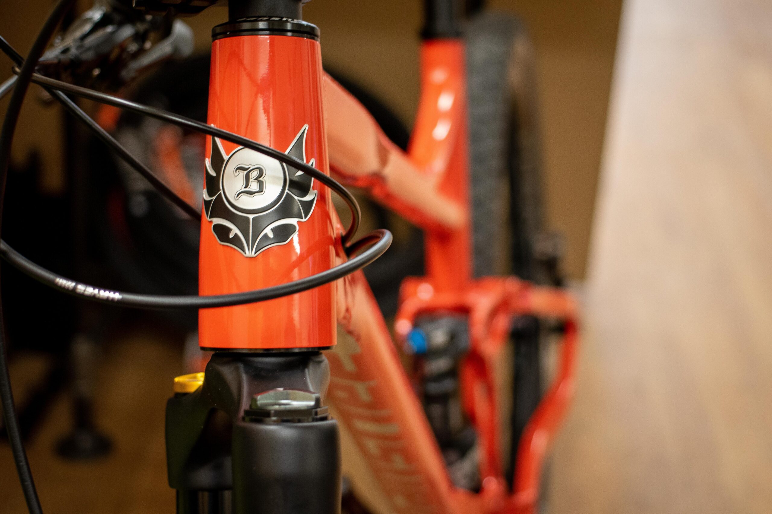Close-up view of a bright orange bicycle frame featuring a logo with a stylized "B" and decorative design on the head tube. Bicycle cables are visible running alongside the frame, with part of the bike's front suspension fork in focus. A blurred background suggests a workshop or storage area.