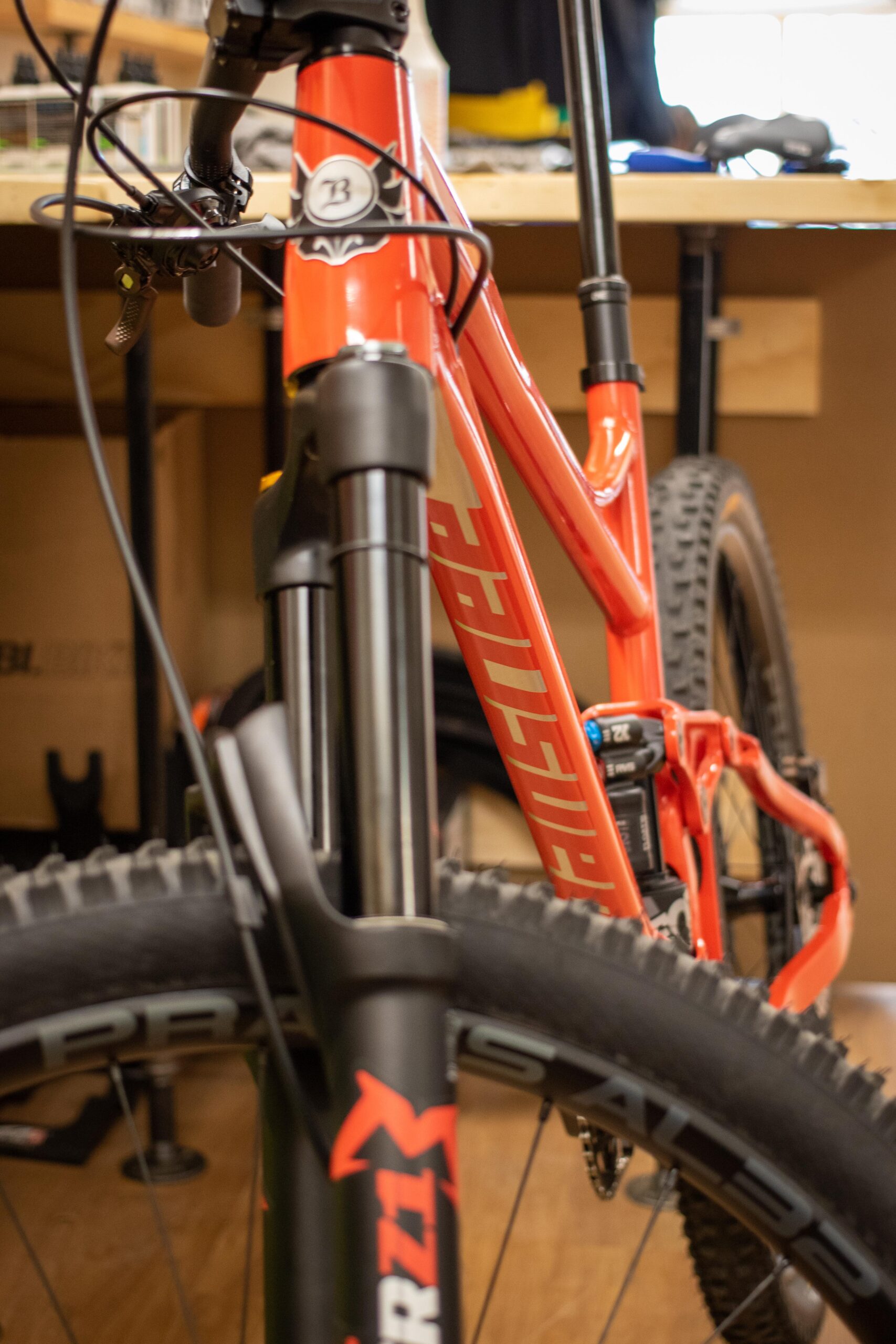 Close-up view of a mountain bike, featuring an orange frame with visible branding, front suspension fork, and a black wheel with a prominent tread pattern. The bike is positioned in a workshop environment with tools and equipment in the background.