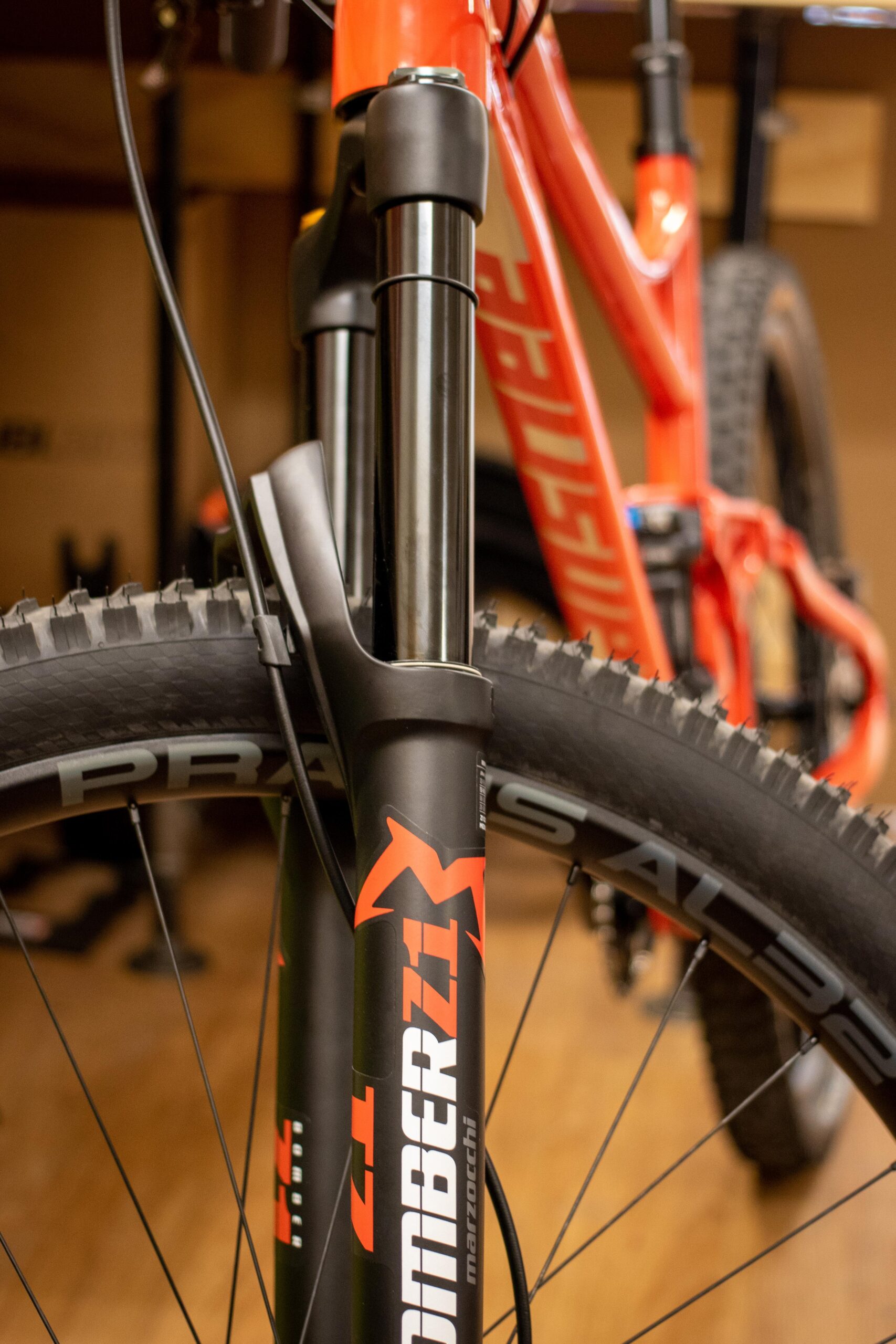Close-up image of a mountain bicycle's front fork showcasing the Marzocchi Bomber Z1 suspension components and the tire labeled "Prazza." The background features part of the bike frame and a wooden floor, indicating a workshop or storage area.