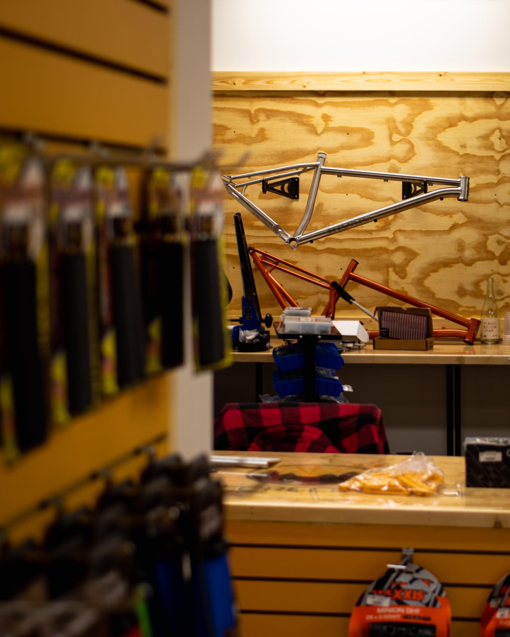 A bicycle workshop containing various bike components and tools. A silver bike frame is mounted on the wall, while additional bike parts are arranged on a workbench in the background. The foreground features a display of bike accessories, including grips and tires, hanging from a wooden panel. A red plaid shirt is draped over a chair, adding a cozy touch to the workspace.