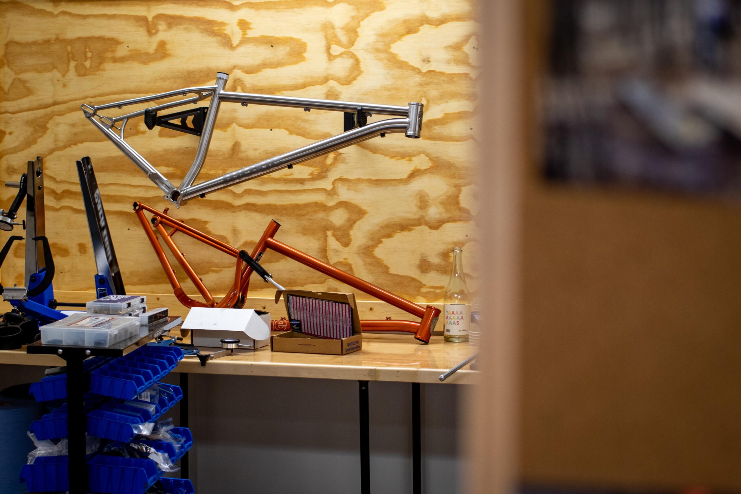 A workspace featuring bike frames in various colors, including silver and orange, mounted on a wooden wall. A table below holds tools and parts organized in blue bins, alongside a box and a bottle. The setting suggests a workshop dedicated to bicycle assembly or maintenance.