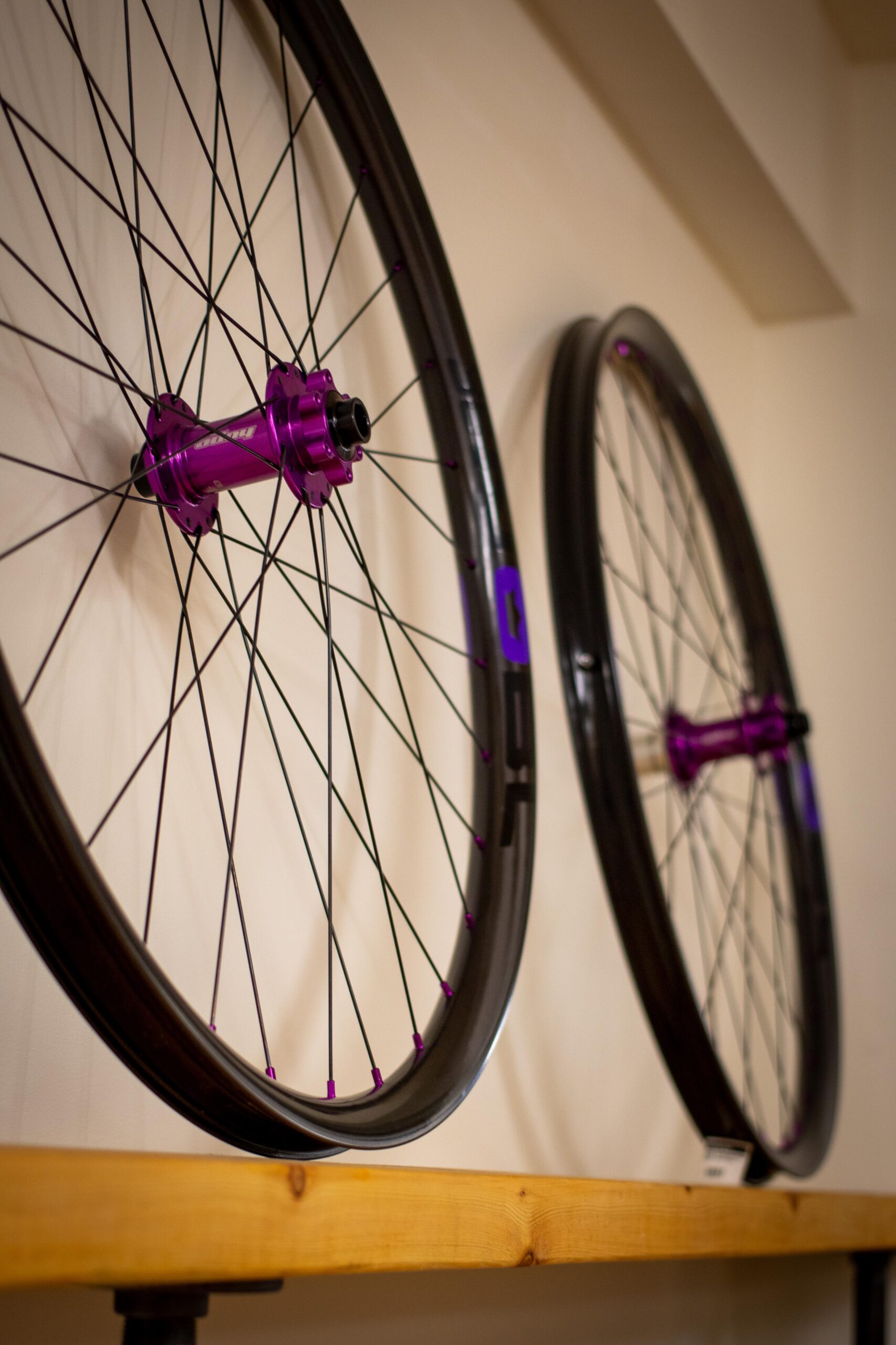 Two bike wheels with black rims and purple hubs are displayed on a wooden shelf against a light-colored wall. The wheels are side-by-side, showcasing their intricate spoke design and vibrant color contrast.