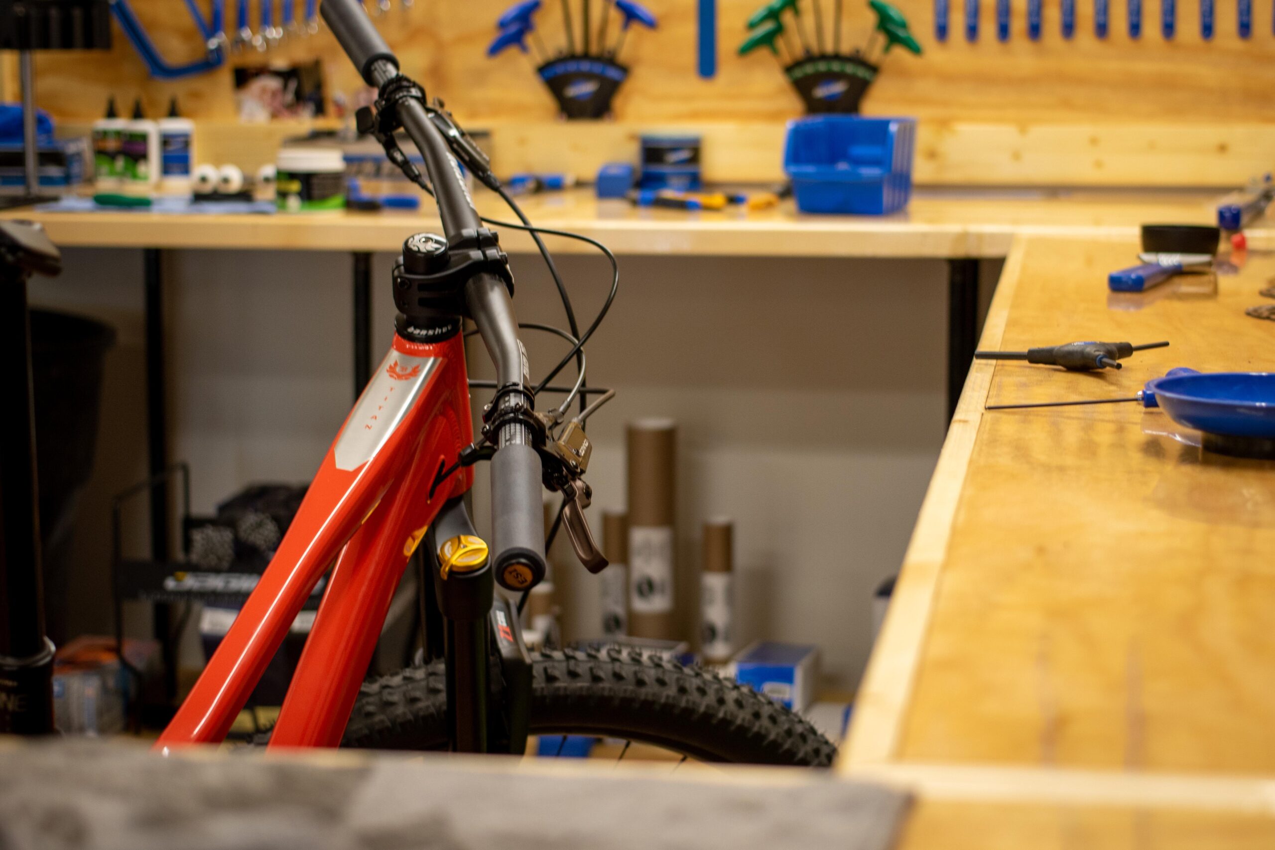 A close-up view of a red bicycle positioned in a workshop setting, with a wooden workbench in the foreground. Various tools and equipment are visible in the background, including blue-handled tools and containers, creating an organized workspace atmosphere.