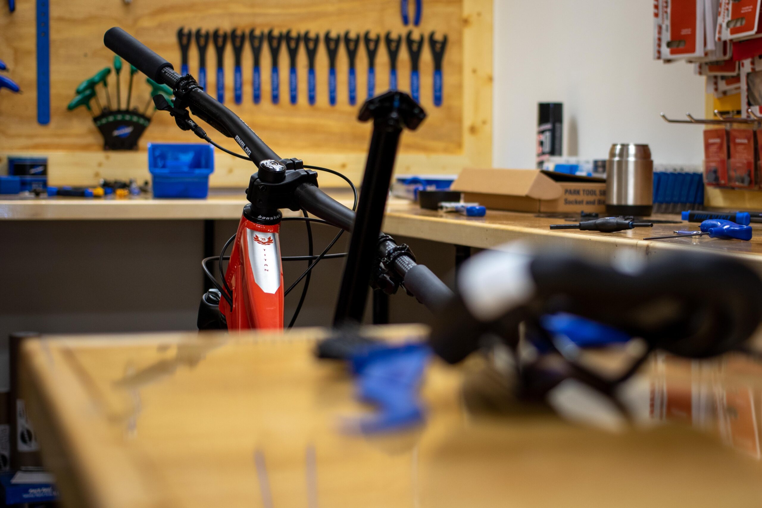 A close-up view of a red mountain bike partially visible in a workshop, with a cluttered workbench in the foreground. The background features a pegboard equipped with various blue and green tools, while the workbench holds additional tools, a coffee mug, and a box.