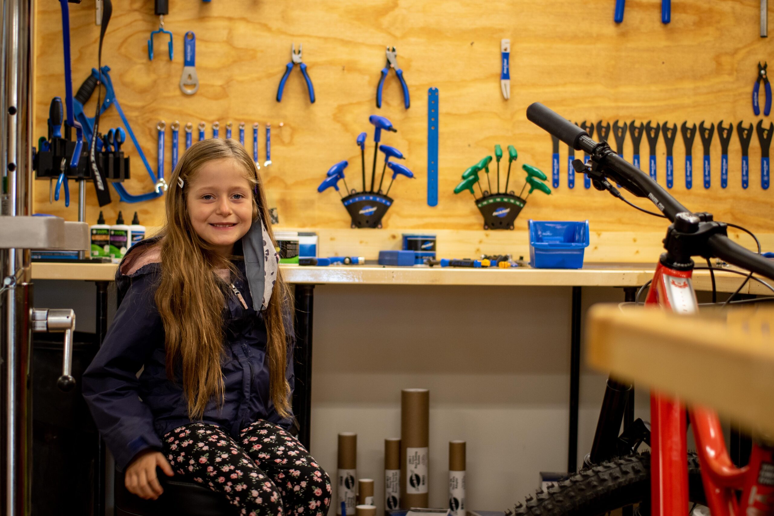 A young girl with long hair smiles while seated on a stool in a workshop. The background features a wooden wall adorned with various hand tools, including wrenches and pliers, as well as a red bicycle partially visible in the frame. The workspace is organized with tools and equipment arranged neatly on a counter.