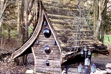 A whimsical, cabin-like structure made of logs with a distinctive sloping roof and round, reflective windows. The building is surrounded by trees, with a small seating area nearby featuring decorative lanterns. The scene evokes a fairytale setting in a wooded area. Columbia Forest Vista Hills mountain bike trail.