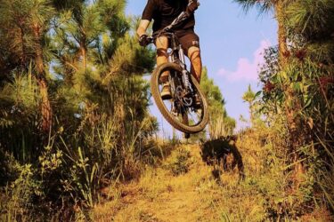 A mountain biker in a black shirt and shorts is jumping off a dirt trail surrounded by tall green pine trees, with blue skies and soft clouds in the background. The biker is airborne with both tires off the ground, showcasing an action-packed moment in outdoor biking. Burning Love MTB Flow Trail mountain bike trail.
