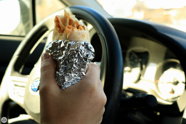 A hand holding a burrito wrapped in foil, resting on the steering wheel of a car, with the interior dashboard visible in the background.
