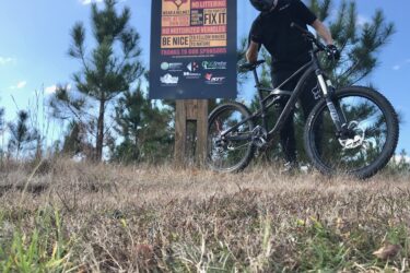 A mountain biker in a black helmet and shirt stands next to a large sign for the "BurningLove MTB Flow Trail." The sign displays rules for trail use and is surrounded by grass and trees under a clear blue sky. Burning Love MTB Flow Trail mountain bike trail.