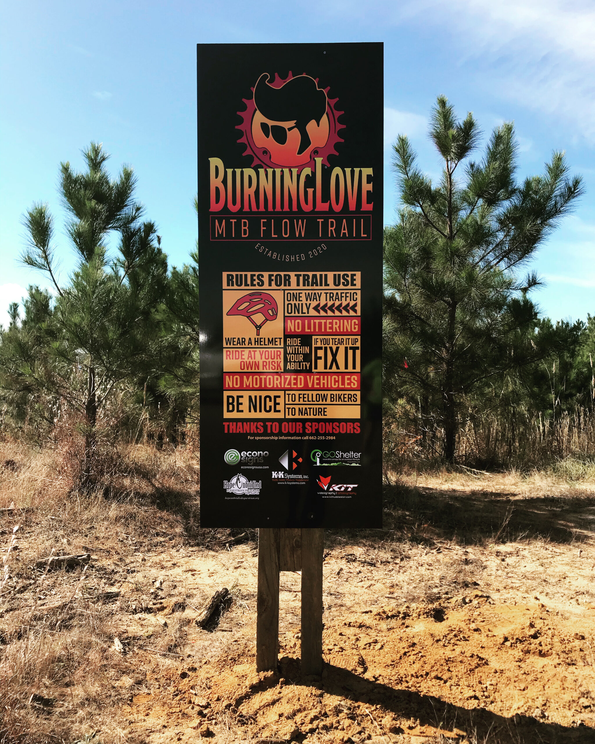 A sign for the Burning Love MTB Flow Trail, featuring rules for trail use such as "One way traffic only," "Wear a helmet," "No littering," and "No motorized vehicles." The sign includes a graphic with a gear and character design, established in 2020, and thanks sponsors at the bottom. The background shows a natural setting with pine trees and a clear sky. Burning Love MTB Flow Trail mountain bike trail.