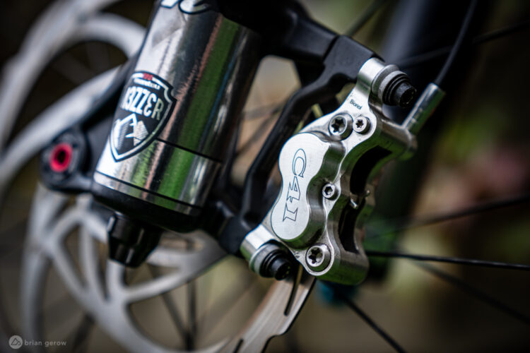 Close-up image of a bicycle's hydraulic disc brake system, featuring a shiny metal brake caliper with the engraving "C41" and a visible brake rotor. A portion of a suspension shock is also visible, displaying a logo. The background is softly blurred, emphasizing the bike components.