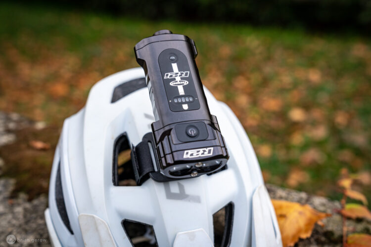 A close-up image of a white bicycle helmet placed on a stone surface, featuring a black bike light mounted on top. The background showcases autumn foliage with blurred colors, indicating an outdoor setting.