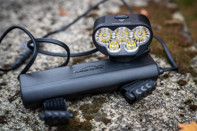 A close-up image of a Magicshine bicycle light system, featuring a compact LED light with four bulbs attached to a rectangular battery pack. The light is connected by a cable, and there are adjustable straps for mounting. The setup is resting on a textured stone surface with a natural background.
