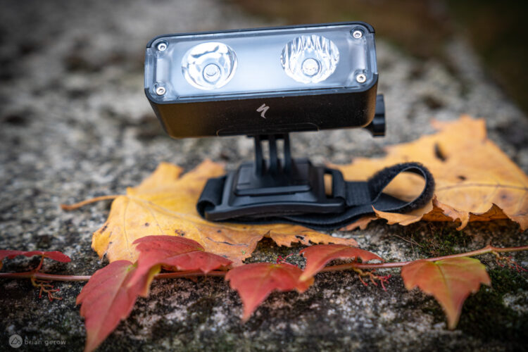 A close-up image of a black bicycle headlight resting on a stone surface, surrounded by autumn leaves. The light features two bright bulbs and is attached to a mounting bracket with a strap. The scene captures the transition of seasons with vibrant red and yellow leaves nearby.