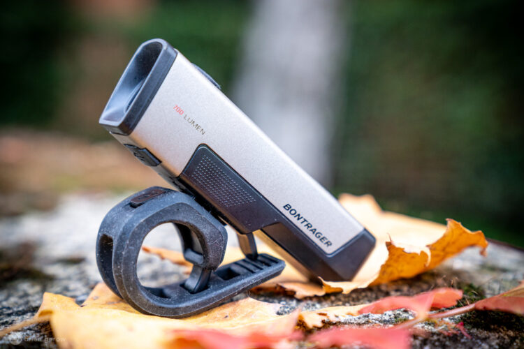 A Bontrager bike light positioned on a fallen leaf, showcasing its sleek silver design, with a label indicating "700 Lumens." The light is mounted on a black rubber holder, emphasizing its utility for cycling. The background features soft, blurred natural elements, hinting at an outdoor setting.