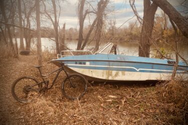 A black mountain bike leaning against a weathered blue boat, partially submerged in a natural setting with bare trees and fallen leaves. The scene reflects a tranquil riverside environment under a cloudy sky. Three Forks Trail mountain bike trail.